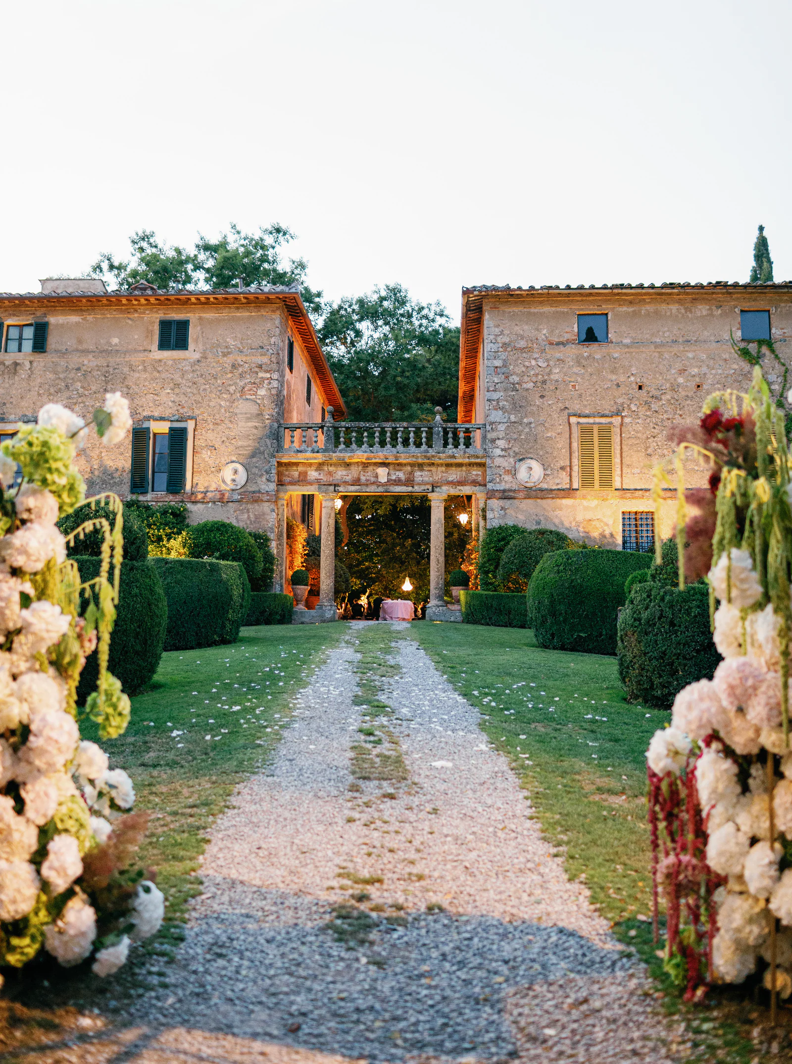 Façade of Borgo Stomennano at dusk, the gravel aisle lined with crimson and white florals, Chianti