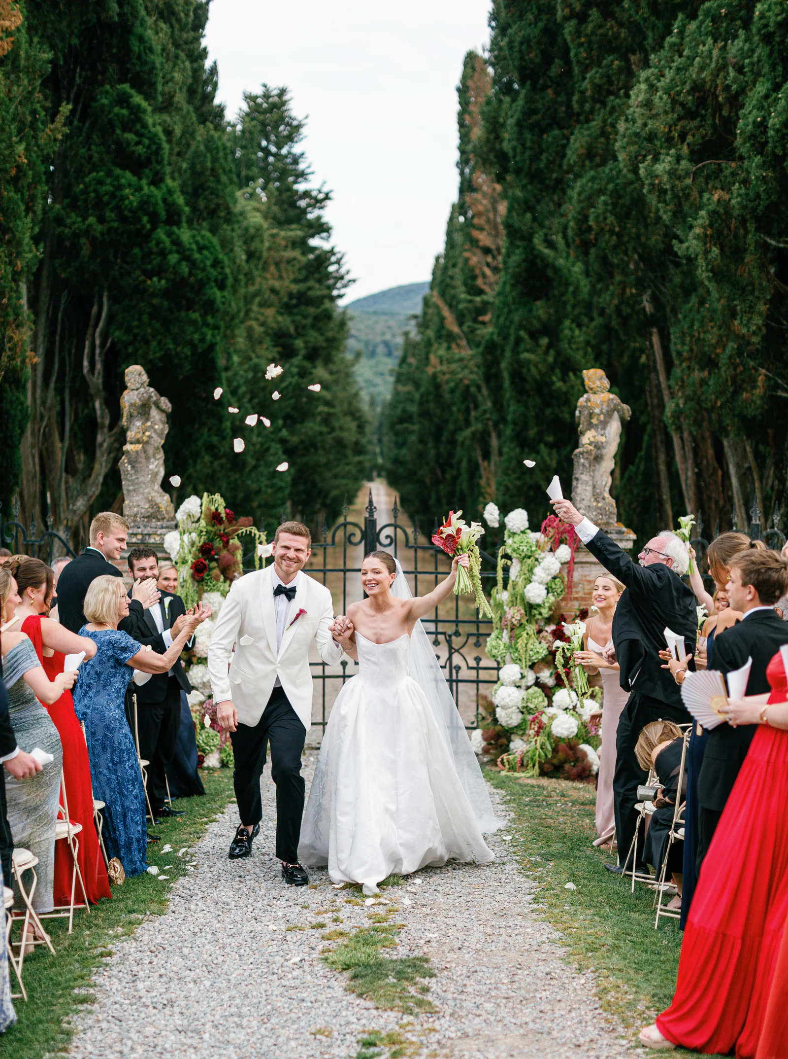 The couple walking back up the aisle between cheering guests along the statue-lined cypress avenue at Borgo Stomennano, Tuscany