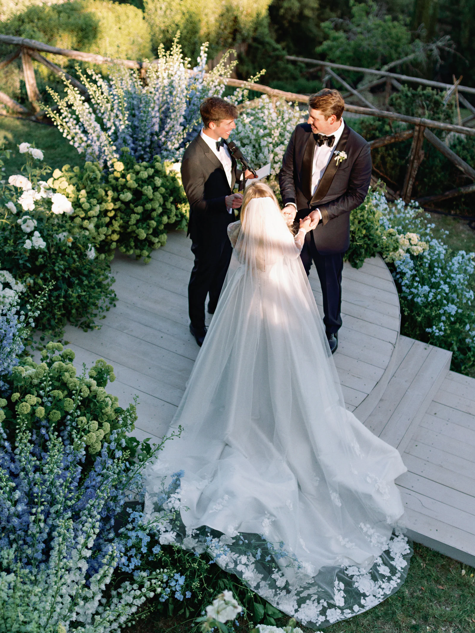Sophie and Gordon holding hands at the ceremony, her veil and lace train across the platform, framed by blue delphiniums and white roses at Castiglion del Bosco, A Rosewood Hotel, Tuscany