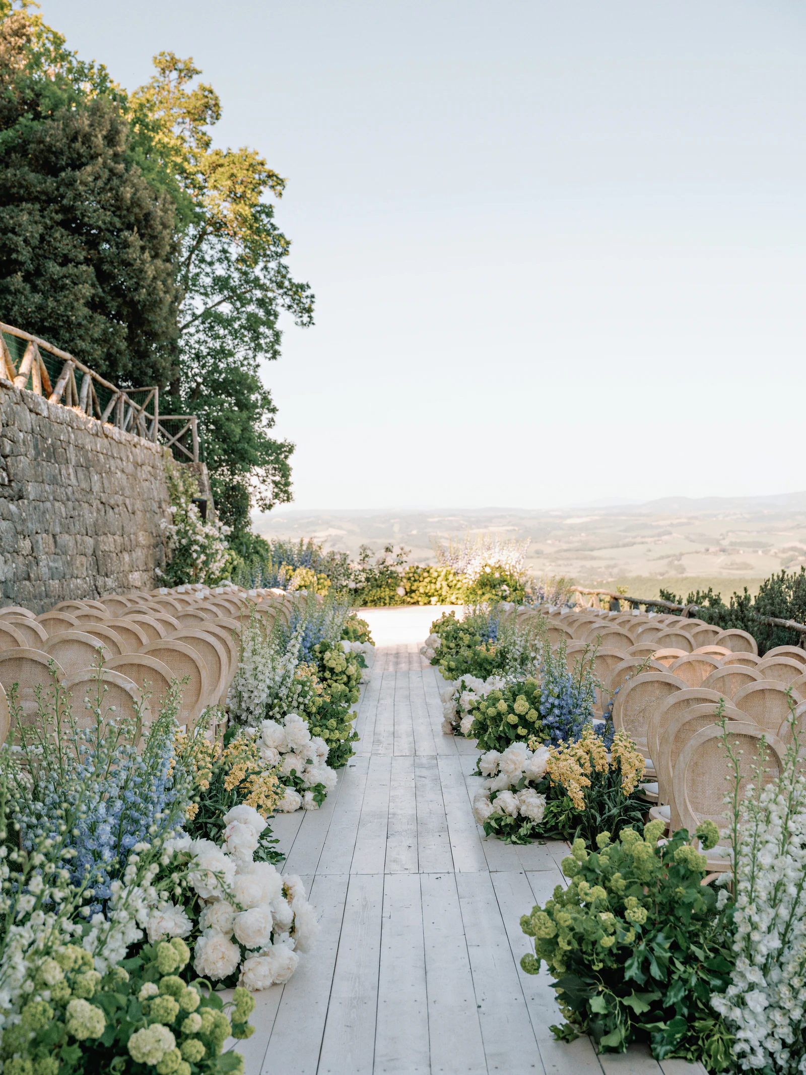 Blue delphiniums and green viburnum at the end of a row of cane chairs, against the stone wall of Castiglion del Bosco, A Rosewood Hotel, Tuscany