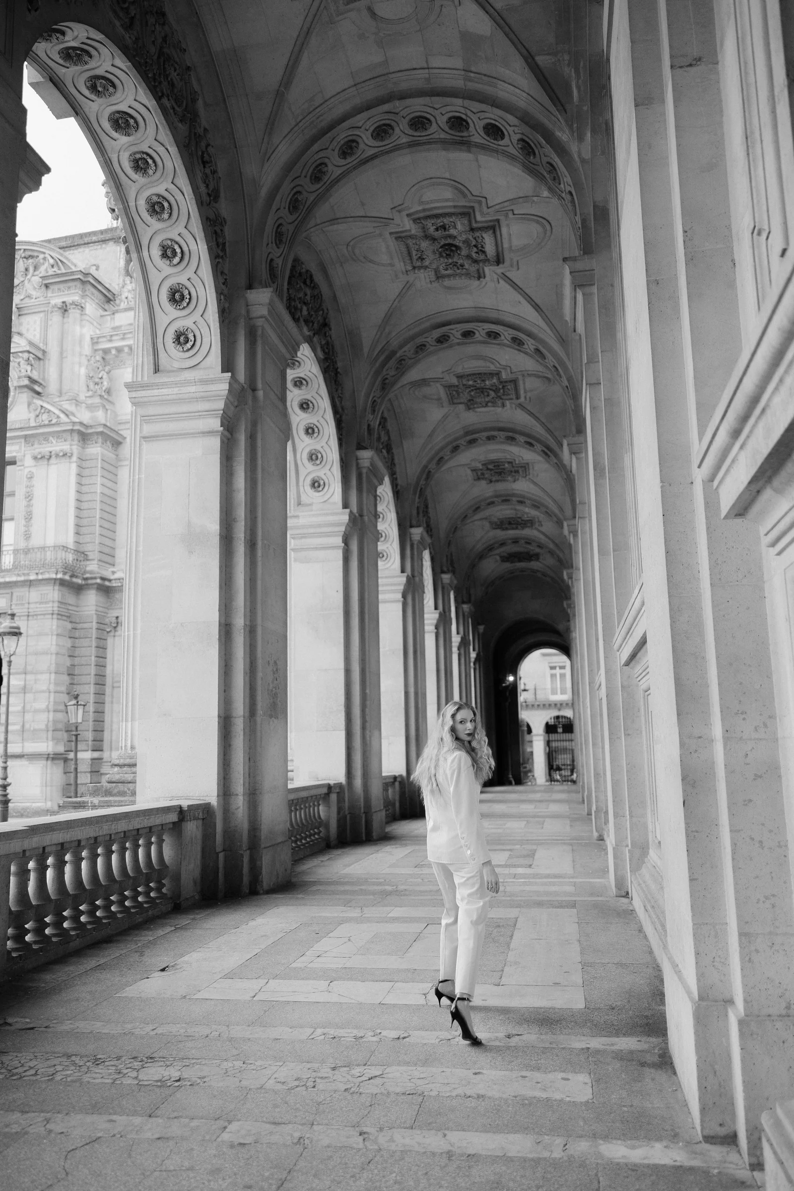 The bride in an ivory trouser suit under an arcade of the Louvre, black and white, Paris