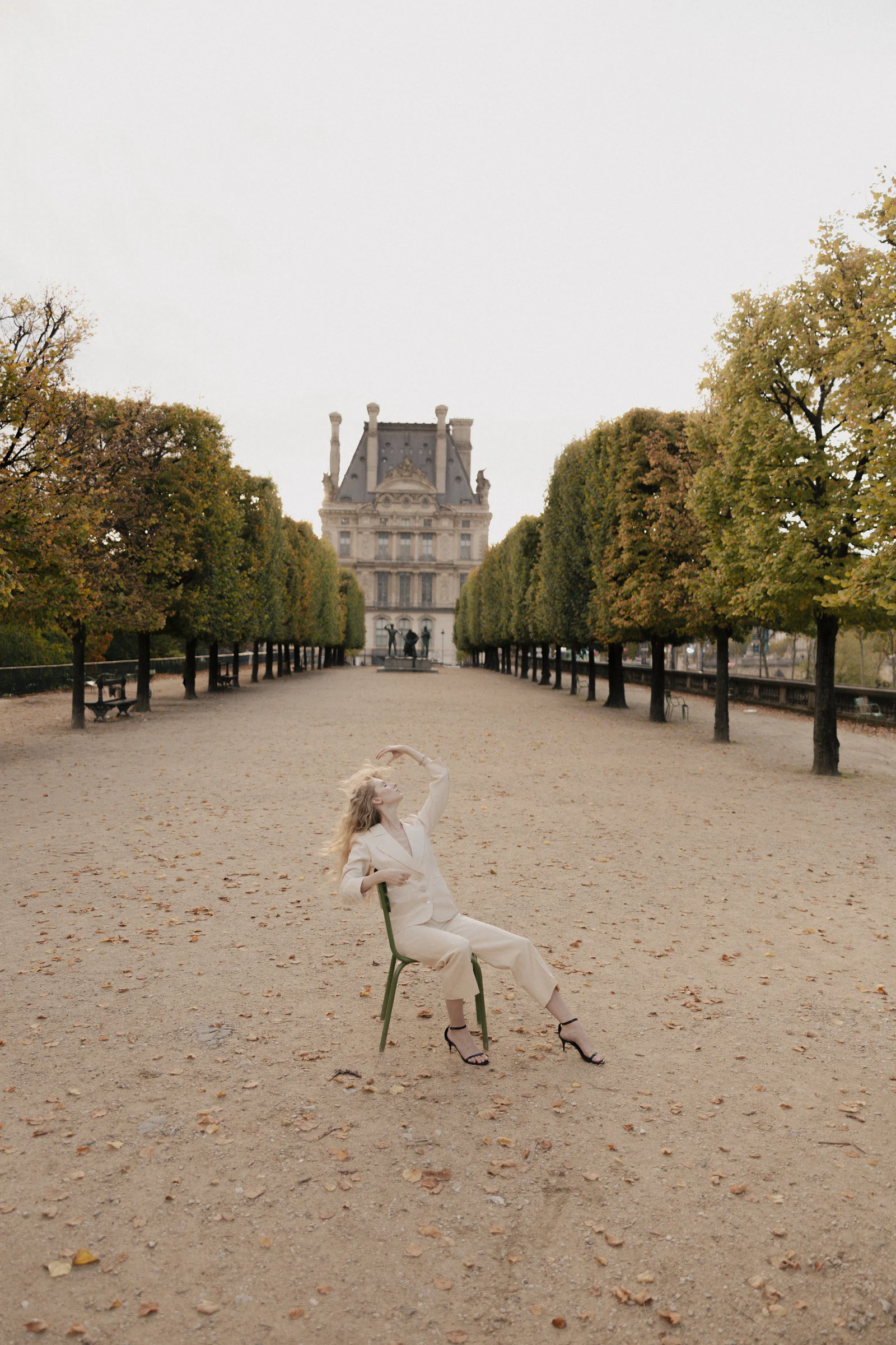 Bride in an ivory trouser suit seated on a green Tuileries chair in the gravel allée, the Louvre beyond, Paris